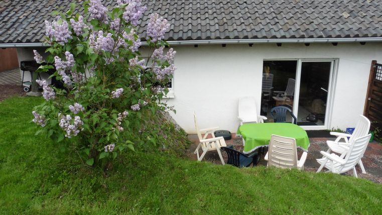 Ein Blick auf ein Haus mit einem Garten. Im Vordergrund steht ein Fliederbaum und es sind mehrere Gartenstühle um einen Tisch gruppiert.