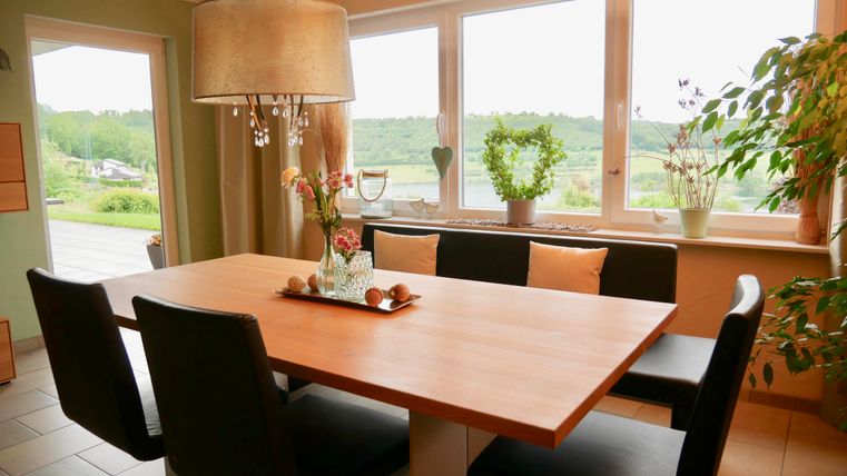 A bright dining room with a large wooden table and black chairs. Through the windows, you can see a green landscape with plants and decorations in the room.
