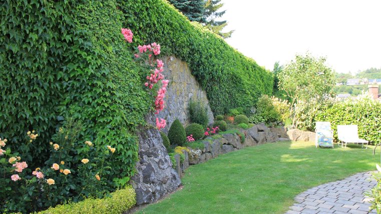 Ein schöner Garten mit grünen Hecken und bunten Blumen. Der Rasen ist gepflegt und es gibt einen gepflasterten Weg.