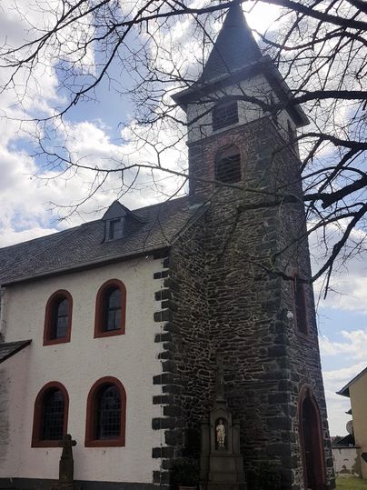 Eine kleine Kirche mit einem steinernen Turm und roten Fensterrahmungen. Der Himmel ist bewölkt, und ein Baum steht im Vordergrund.