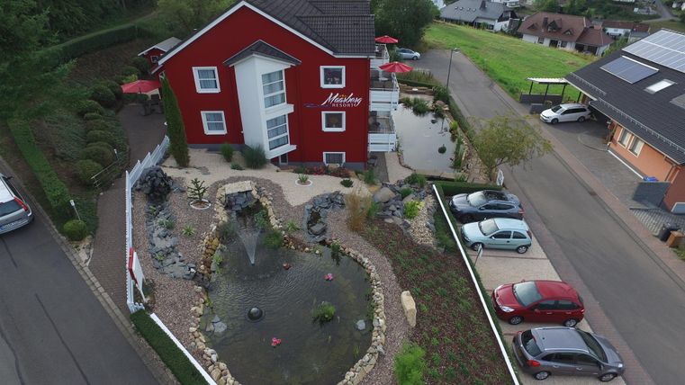 Ein rotes Haus mit einem schönen Garten und einem Teich. Parkplätze sind vor dem Gebäude vorhanden.
