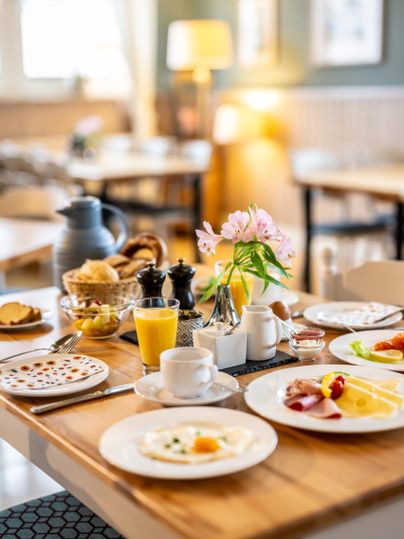 A hearty breakfast on a wooden table with fresh rolls, fruit, and drinks. A pretty flower in a vase creates a welcoming atmosphere.
