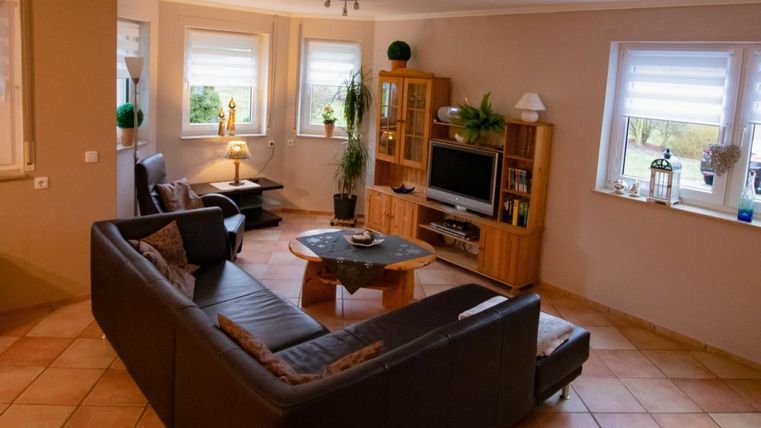 A cozy living room with a large, dark couch and a table in the middle. There are windows with light and a wooden dresser that houses a television.