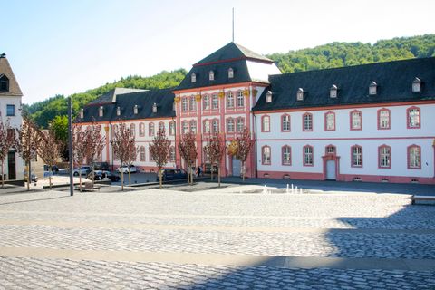 A historic building with red and white masonry stands in a square. In the background, green hills and a clear sky can be seen.