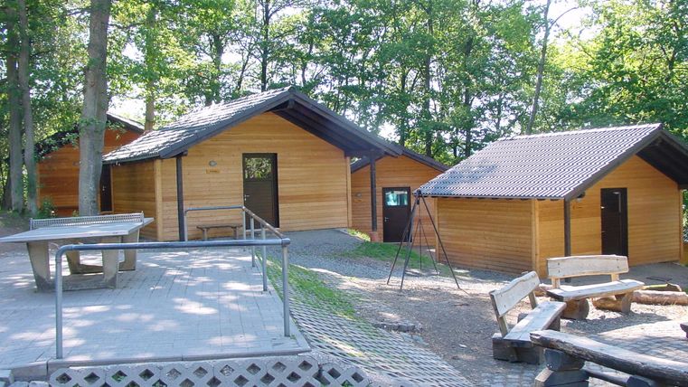 Drie houten hutten in het bos, omringd door bomen. Een pingpongtafel en houten banken staan op een verharde plek.