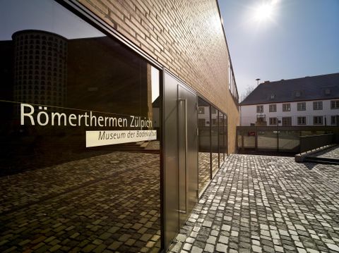 Ein modernes Gebäude mit einer großen Glasfront, das die Römerthermen in Zülpich beherbergt. Im Hintergrund sind traditionelle deutsche Häuser zu sehen.