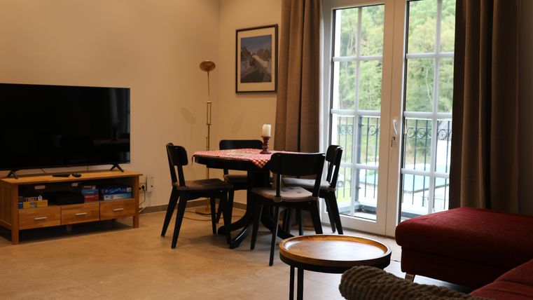 A modern living room with a television, a dining table, and chairs. Large windows let in a lot of light and offer a view of the greenery.