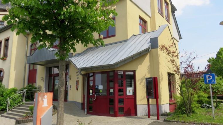 Ein modernes Gebäudeeingang mit rot-weißen Fenstern und einem übersichtlichen Eingangsbereich. Vor dem Gebäude stehen einige Bäume und ein Parkplatz ist in der Nähe.