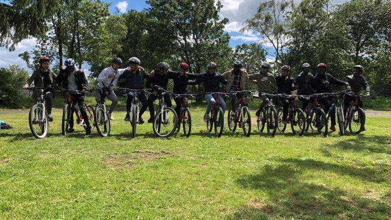Eine Gruppe von zehn Radfahrern steht auf Fahrrädern auf einer grünen Wiese. Im Hintergrund sind Bäume und ein blauer Himmel mit einigen Wolken zu sehen.