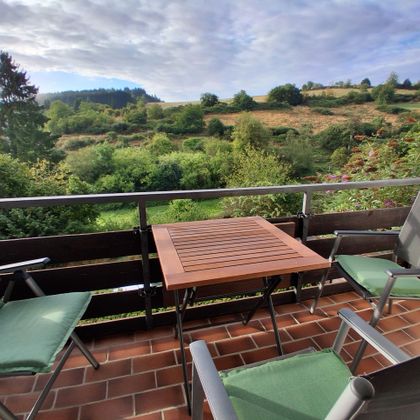 Ein schöner Balkon mit zwei Stühlen und einem Holztisch. Im Hintergrund sind grüne Hügel und eine bewölkte, aber helle Himmel zu sehen.