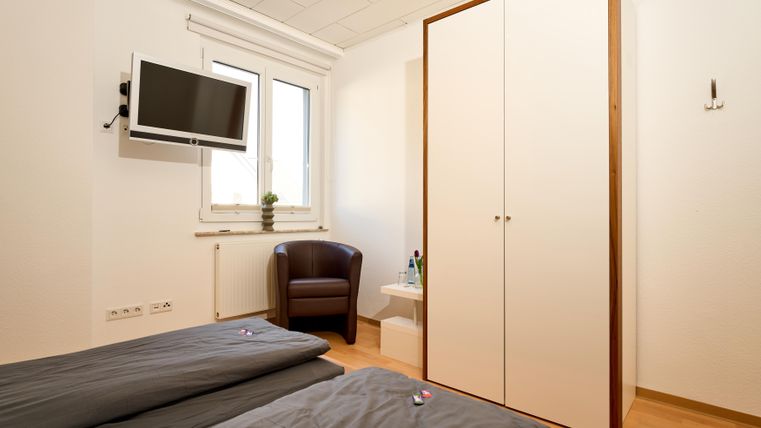 Ein einfaches Schlafzimmer mit zwei Matratzen, einem Fernseher und einem Kleiderschrank. Ein bequemer Sessel und ein Tisch mit dekorativen Gegenständen ergänzen den Raum.