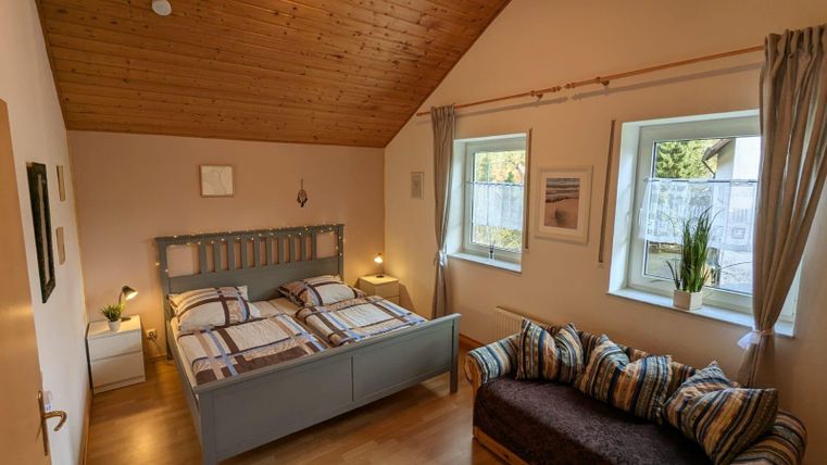 A cozy bedroom with a double bed and a sofa. The walls are light, and large windows let in plenty of light.