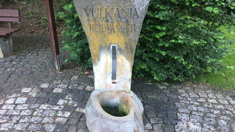 A stone spring fountain with the inscription "Vulkana Healing Water". The fountain is surrounded by green plants and stands on a paved path.