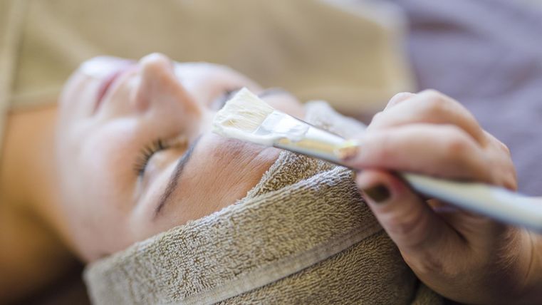 Eine Person erhält eine Gesichtsbehandlung. Sie liegt entspannt mit einem Handtuch um den Kopf, während eine andere Person Produkte aufträgt.