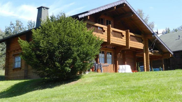 Ein gemütliches Holzhaus steht auf einer Wiese, umgeben von Bäumen. Der Himmel ist klar und sonnig.