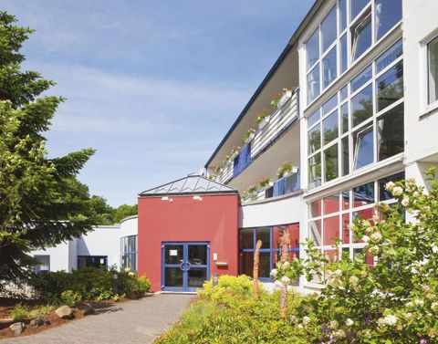 A modern building with large windows and a red entrance. Surrounded by lush greenery and colorful flowers.