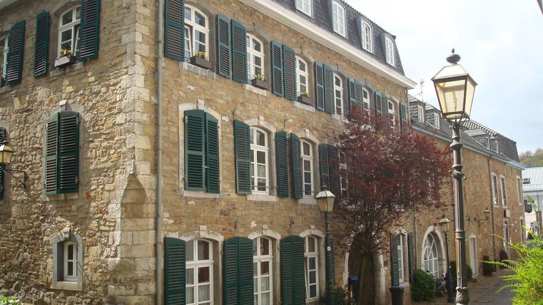 Een historisch gebouw met een stenen gevel en groene luiken. Op de voorgrond loopt een geplaveide straat en er zijn lantaarns te zien.