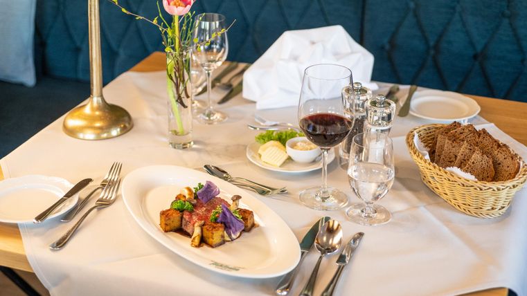 Ein schön gedeckter Tisch mit einem eleganten Essen in der Mitte. Daneben stehen Gläser mit Wasser und Wein sowie ein Korb mit Brot.