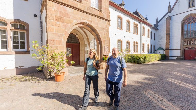 Two people are walking in a historic courtyard with beautiful stone architecture. The sun is shining and there are green plants in the foreground.