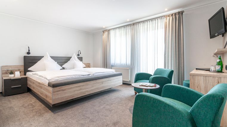 Ein modernes Hotelzimmer mit einem großen Bett und zwei grünen Sesseln. Die Fenster lassen viel Tageslicht herein und der Raum wirkt hell und einladend.