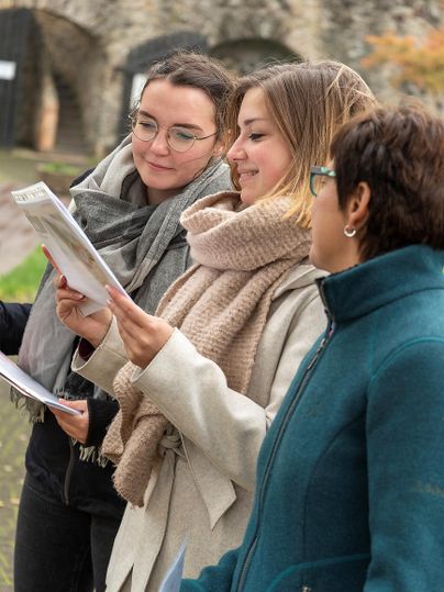 Three women outdoors, dressed warmly, are together looking at a document. In the background, old stone walls can be seen.