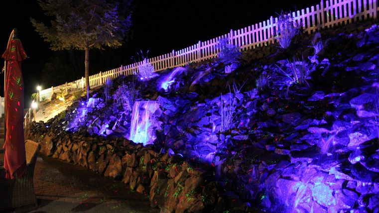 Ein schöner, beleuchteter Steinwall bei Nacht mit violetten Lichtern. Im Hintergrund ist ein heller Zaun und einige Pflanzen sichtbar.