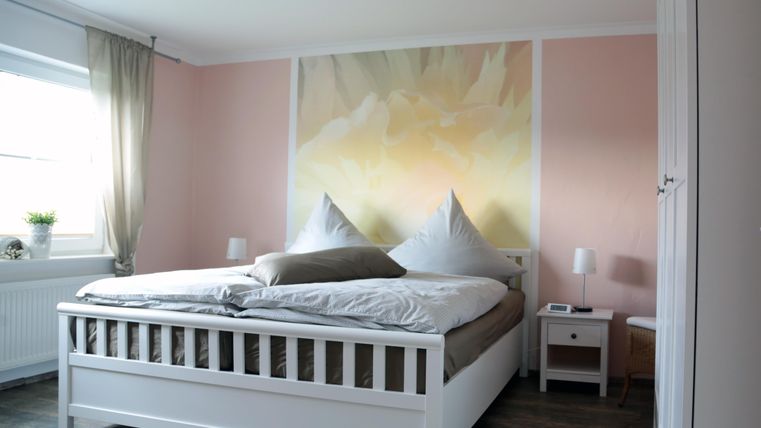 Ein helles Schlafzimmer mit einem großen Doppelbett und einer blumigen Wandgestaltung. Neben dem Bett stehen zwei Nachttische und ein Fenster mit Vorhängen.