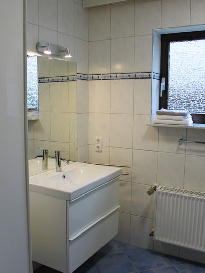 Een moderne badkamer met een dubbele wastafel en een grote spiegel. De wanden zijn licht betegeld, en een raam zorgt voor natuurlijk licht.