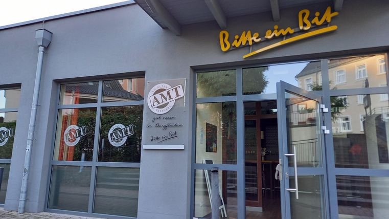 Ein modernes Gebäude mit großen Fenstern und einer einladenden Eingangstür. Das Schild zeigt den Namen "Bitte ein Bit" sowie das Logo "AMT".