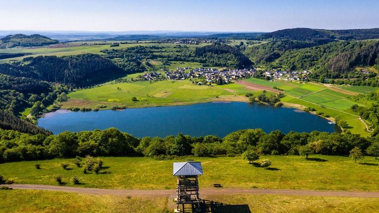 A picturesque landscape with a lake and green meadows. In the background, there are gentle hills and a small village.