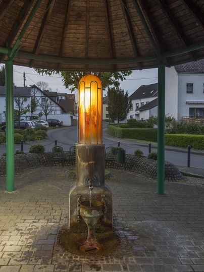 Ein historischer Brunnen in einem Pavillon, beleuchtet von oben. Umgeben von einer ruhigen Straße und Wohnhäusern.