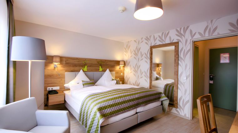 Ein modernes Hotelzimmer mit einem Doppelbett, einem gemütlichen Sessel und einer eleganten Beleuchtung. Die Wände sind geschmackvoll dekoriert und ein großer Spiegel sorgt für ein helles Ambiente.