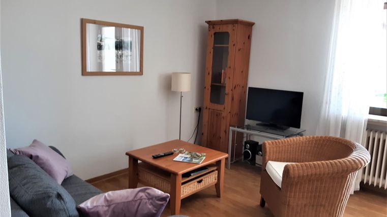 Een gezellige woonkamer met een bank, een fauteuil en een houten tafel. Er is een televisie en veel licht door het raam.