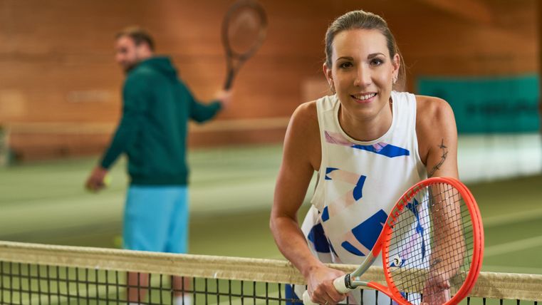 Eine sportliche Frau posiert am Tennisnetz mit einem Schläger in der Hand. Im Hintergrund spielt ein Mann Tennis.