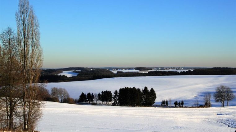 Een besneeuwd landschap met een wijd uitzicht over velden en bomen. De lucht is helder en blauw.