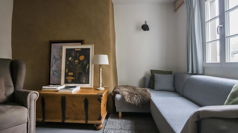 Ein gemütlicher Wohnraum mit einem grauen Sofa, einem Holzkaffee-Tisch und einem bequemen Sessel. An der Wand hängen einige Bilder, und das Licht fällt sanft durch das Fenster.