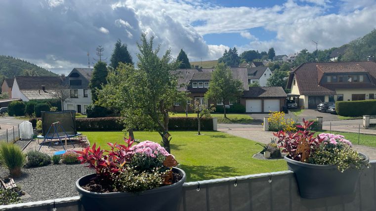 Ein schöner Ausblick auf eine Wohnsiedlung mit gepflegten Gärten und einem blauen Himmel. Im Vordergrund stehen blühende Pflanzen in Kästen.