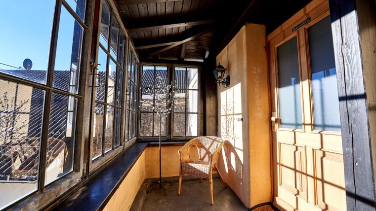 A bright balcony with large windows and a wooden table. A single chair stands by the door and it seems that the sun is shining.