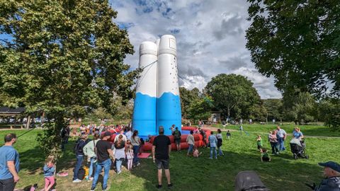 Een grote, kleurrijke sculptuur in de vorm van twee melktanks staat in het park. Veel mensen, waaronder kinderen, verzamelen zich eromheen en genieten van de zonnige dag.