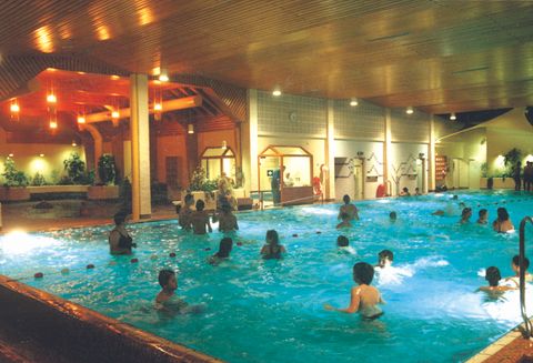 Een binnenzwembad met veel mensen die in het water zwemmen en spelen. De omgeving is helder verlicht en heeft een ontspannen sfeer.