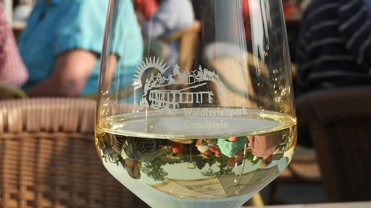 Weinglas mit Gravur "Waldferienpark Gerolstein" auf Tisch, unscharfe Personen im Hintergrund.