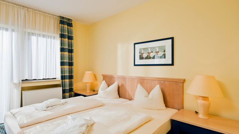 Ein gemütliches Schlafzimmer mit einem Doppelbett und hellen Farben. Es gibt einen Schreibtisch, Lampen und ein Fenster mit Vorhängen.