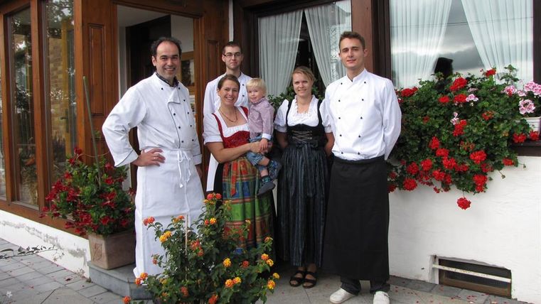 Eine Gruppe von fünf Personen steht vor einem Restaurant mit blühenden Blumen. Die Menschen tragen traditionelle Trachten und Kochuniformen.