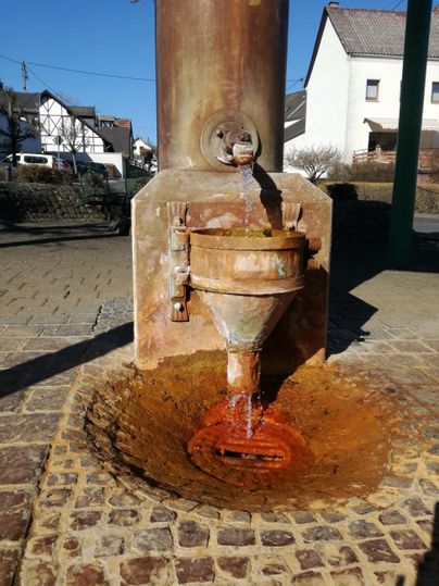 Eine alte Wasserpumpe mit rostigen Teilen, aus der braunes Wasser fließt. Im Hintergrund sind einige Gebäude zu sehen.