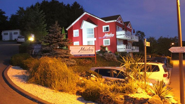 Ein rotes Gebäude mit mehreren Balkonen steht in der Dämmerung. Umgeben von beleuchteten Pflanzen und Fahrzeugen, strahlt es eine gemütliche Atmosphäre aus.