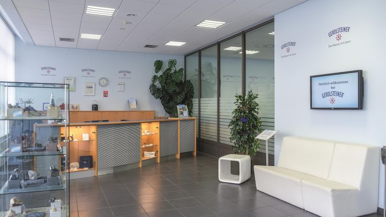 Modern visitor center with reception desk, glass showcase, white benches, and plants. Gerolsteiner logos are visible on the walls.