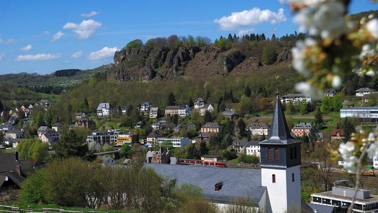 Panorama von Gerolstein mit Kirche im Vordergrund, umgeben von Häusern und bewaldeten Hügeln unter blauem Himmel.