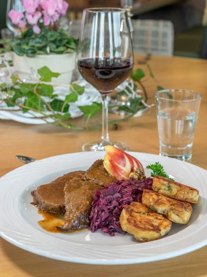 A plate with tender meat, red cabbage, and potato dumplings. In the background, there is a glass of red wine and a glass of water.
