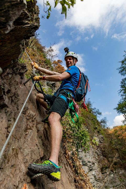 Ein Mann klettert an einer Felswand und trägt einen Helm sowie Kletterausrüstung. Der Himmel ist blau mit einigen Wolken und die Umgebung ist grün.