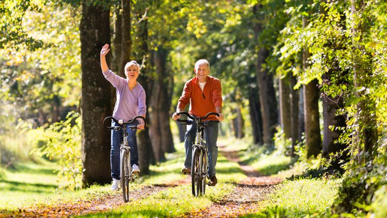 Ein älteres Paar fährt fröhlich mit dem Fahrrad auf einem Waldweg. Umgeben von Bäumen genießen sie die Natur.